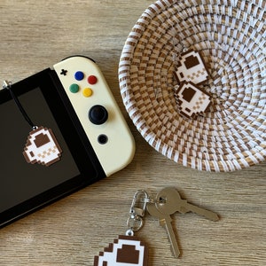 May include: A set of three pixelated coffee cup charms. The charms are made of felt and are in the shape of a coffee cup with a brown liquid inside. The charms are attached to a key chain, a pair of earrings, and a Nintendo Switch console.