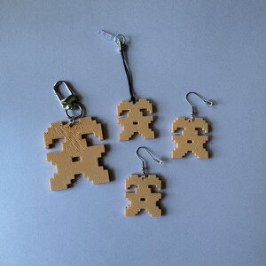 May include: Three tan-colored pixelated figures, each with a different style of attachment. The figure on the left is attached to a silver key chain, the figure in the middle is attached to a black string, and the figure on the right is attached to a silver earring hook.