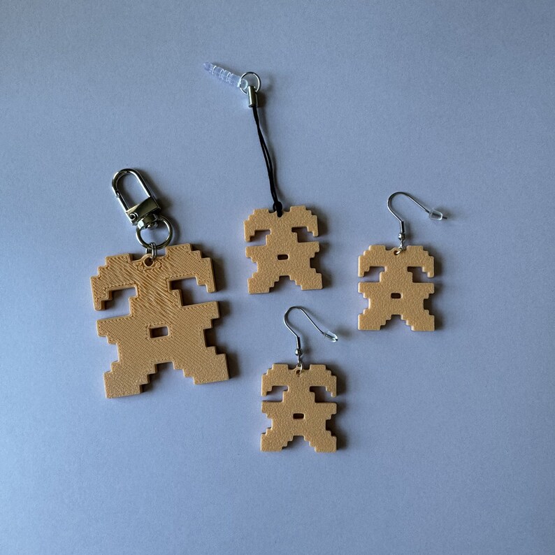 May include: Three tan-colored pixelated figures, each with a different style of attachment. The figure on the left is attached to a silver key chain, the figure in the middle is attached to a black string, and the figure on the right is attached to a silver earring hook.