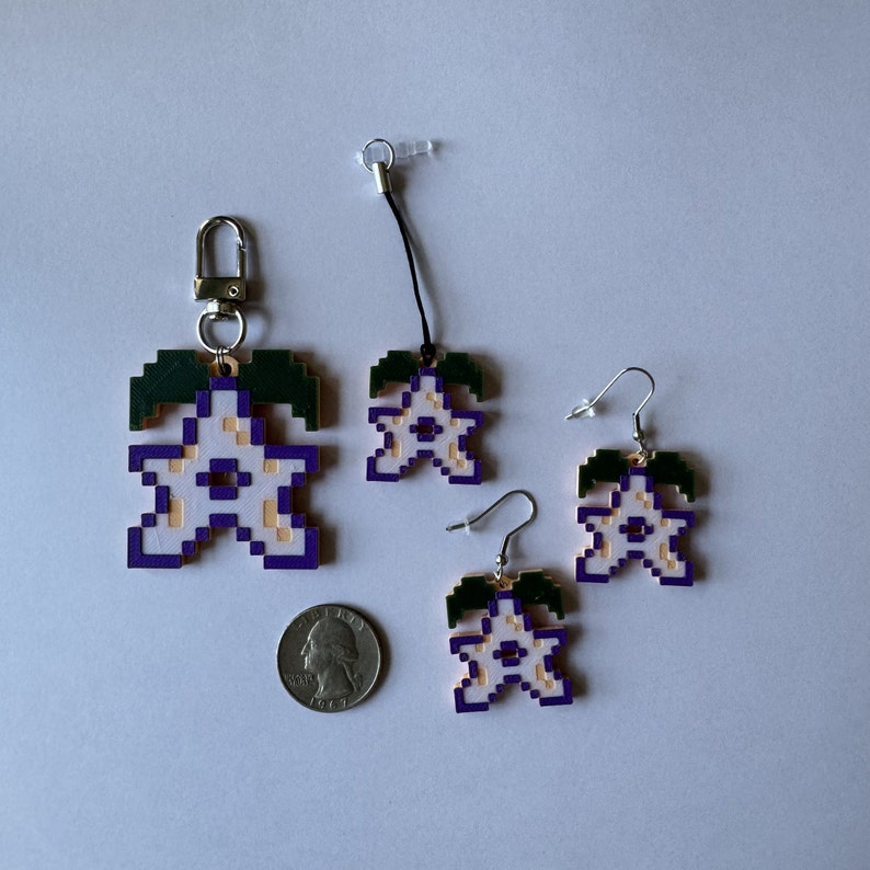 May include: Three purple and white flower charms with gold accents. The charms are in the shape of a five-pointed star with a green stem and leaves. Two of the charms are earrings and one is a keychain.