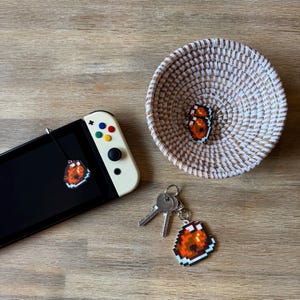 May include: A white and brown woven basket with a red and orange pixelated mushroom charm inside. A Nintendo Switch console with a red and orange pixelated mushroom charm attached to it. A set of keys with a red and orange pixelated mushroom charm attached to it.