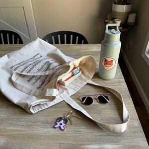 May include: A beige canvas tote bag with musical notes printed on it. The bag is filled with a book and a small box. A pair of pink sunglasses, a key chain with a purple flower design, and a light blue water bottle with a fox sticker are on the table next to the bag. The sticker says "Grey Fox & Co."