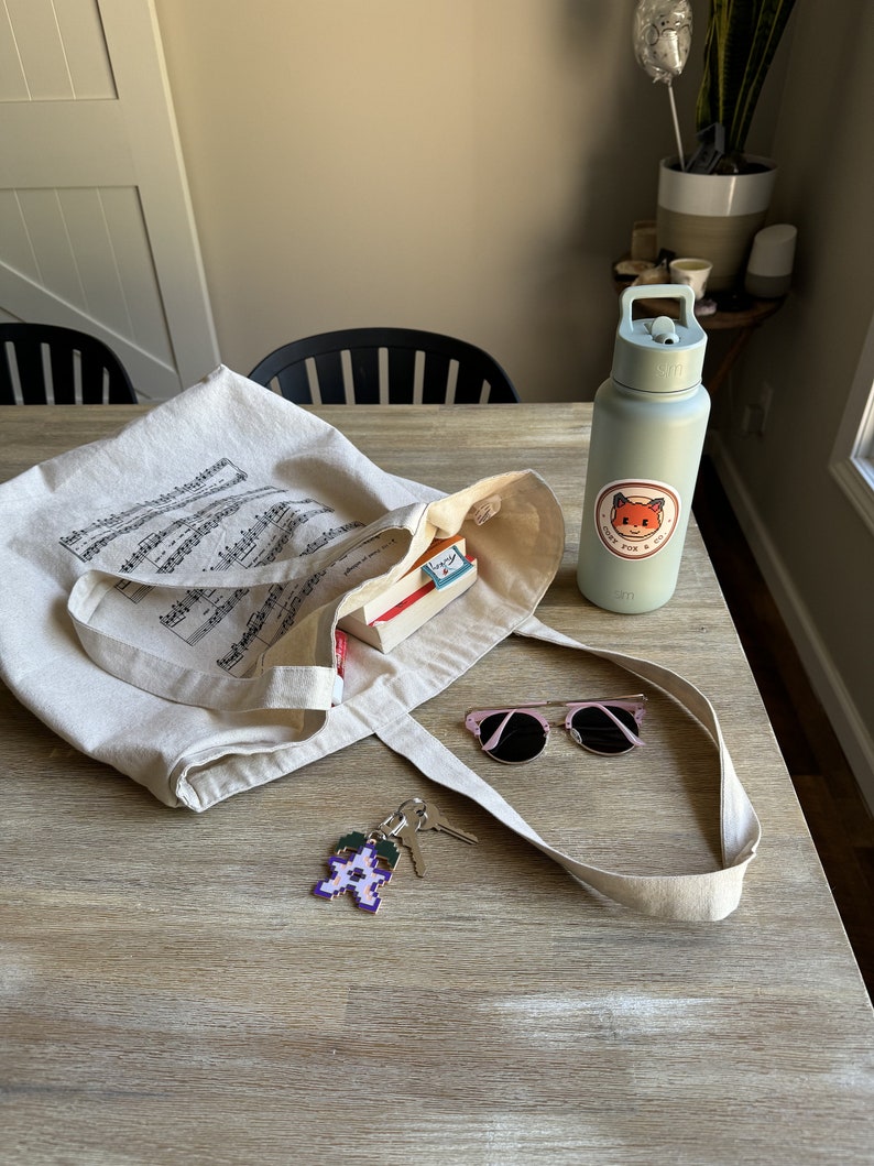 May include: A beige canvas tote bag with musical notes printed on it. The bag is filled with a book and a small box. A pair of pink sunglasses, a key chain with a purple flower design, and a light blue water bottle with a fox sticker are on the table next to the bag. The sticker says "Grey Fox & Co."