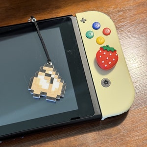 Puede incluir: Una consola Nintendo Switch amarilla con una pantalla negra y un amuleto de personaje pixelado blanco y marrón sujeto a la parte superior. La consola tiene un botón en forma de fresa rojo y blanco en el lado derecho.