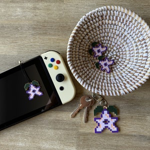 May include: A white and purple pixelated flower charm with green leaves hanging from a keychain. The charm is attached to a set of keys and a Nintendo Switch console.