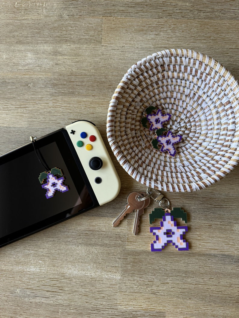 May include: A white and purple pixelated flower charm with green leaves hanging from a keychain. The charm is attached to a set of keys and a Nintendo Switch console.