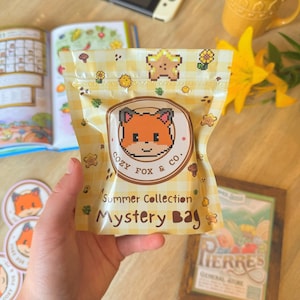 May include: A yellow and white checkered "Mystery Bag" from the "Summer Collection" by "Cozy Fox & Co." features a pixelated fox logo. The bag is held in a hand, with stickers, a mug, a game console, and a book in the background.