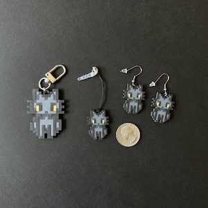 Puede incluir: Un conjunto de accesorios con forma de gato pixelado, que incluye un llavero, un charm para tel&eacute;fono y un par de pendientes. Las figuras de gato grises tienen ojos amarillos y se muestran sobre un fondo oscuro. Se incluye una moneda como referencia de tama&ntilde;o.