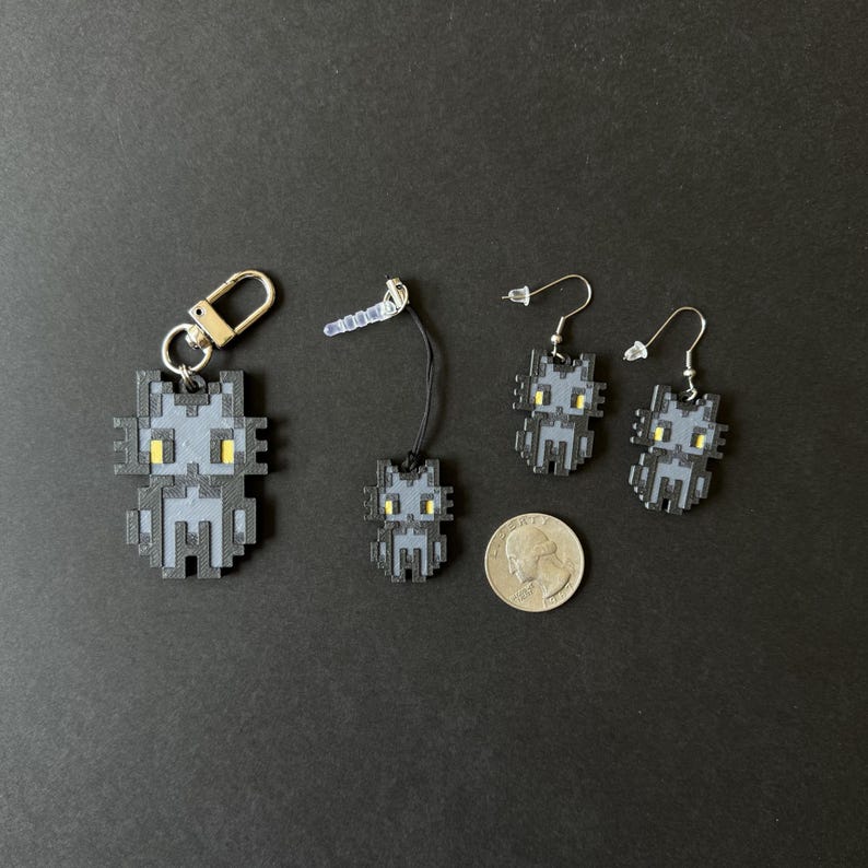 Puede incluir: Un conjunto de accesorios con forma de gato pixelado, que incluye un llavero, un charm para tel&eacute;fono y un par de pendientes. Las figuras de gato grises tienen ojos amarillos y se muestran sobre un fondo oscuro. Se incluye una moneda como referencia de tama&ntilde;o.