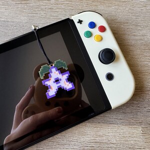 May include: A purple and green pixelated character charm attached to a black lanyard. The charm is hanging from a white Nintendo Switch console.