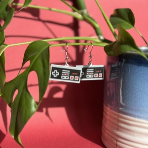 Puede incluir: Un par de pendientes de plata con colgantes de mando de videojuego en blanco y negro. Los colgantes tienen botones rojos y un signo más.