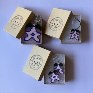 May include: Three small boxes with the text "Cozy Fox & Co." printed on them. Each box contains a different flower-shaped charm with a purple outline and a white center. The charms are made of plastic and are attached to a silver chain or a pair of earrings.