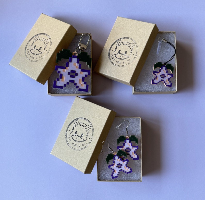 May include: Three small boxes with the text "Cozy Fox & Co." printed on them. Each box contains a different flower-shaped charm with a purple outline and a white center. The charms are made of plastic and are attached to a silver chain or a pair of earrings.