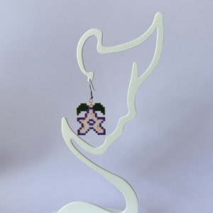 May include: A pair of earrings featuring a pixelated flower design in shades of pink, purple, and green. The flower is hanging from a silver hook.