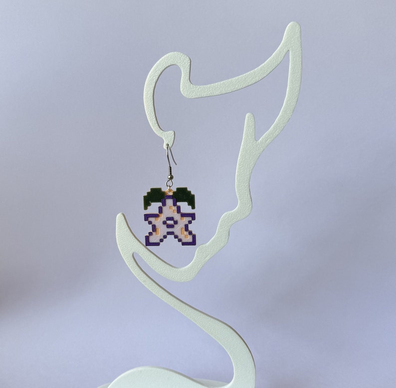 May include: A pair of earrings featuring a pixelated flower design in shades of pink, purple, and green. The flower is hanging from a silver hook.