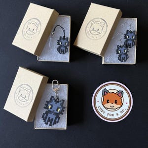 Puede incluir: Varios accesorios con tem&aacute;tica de gato pixelado, incluyendo un colgante, pendientes y un llavero, presentados en peque&ntilde;as cajas de cart&oacute;n. Cada caja presenta un logotipo con una ilustraci&oacute;n de zorro y el texto "COZY FOX & CO.". Tambi&eacute;n se ve una pegatina con el mismo logotipo.