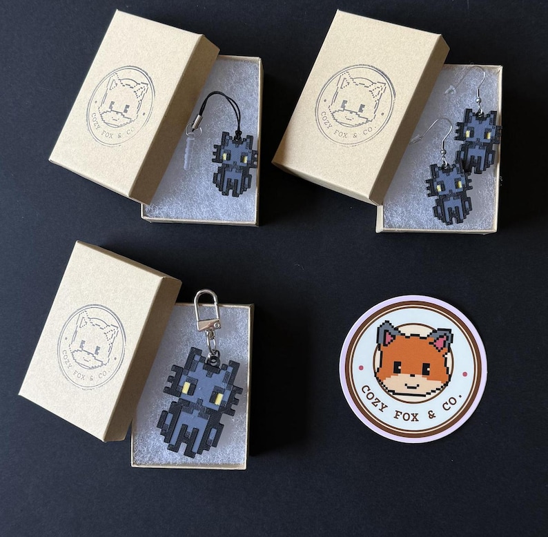 Puede incluir: Varios accesorios con tem&aacute;tica de gato pixelado, incluyendo un colgante, pendientes y un llavero, presentados en peque&ntilde;as cajas de cart&oacute;n. Cada caja presenta un logotipo con una ilustraci&oacute;n de zorro y el texto "COZY FOX & CO.". Tambi&eacute;n se ve una pegatina con el mismo logotipo.