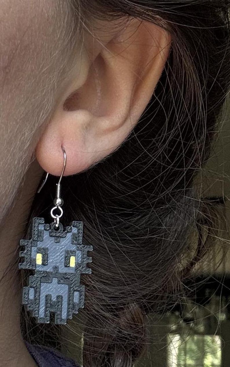 Puede incluir: Pendientes de gato pixelados grises con ojos amarillos. Los pendientes tienen un gancho plateado. Un accesorio &uacute;nico para los amantes de los gatos. Una adici&oacute;n divertida y peculiar a cualquier atuendo.