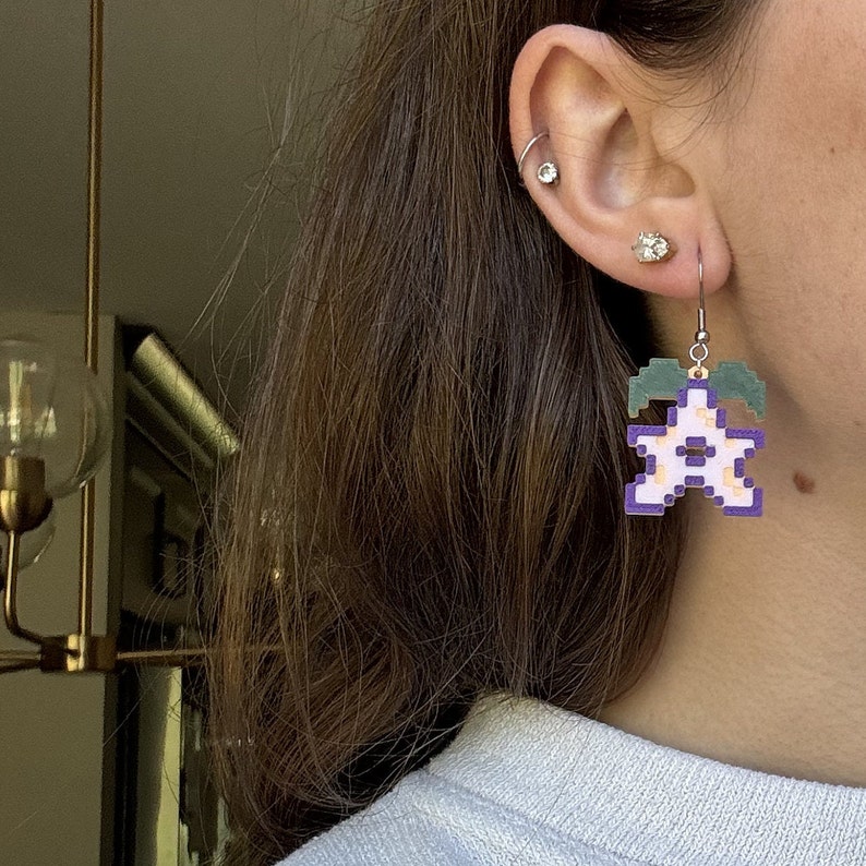 May include: Purple and green pixelated flower earring with a white center. The earring is hanging from a silver hook.