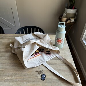 Puede incluir: Una bolsa tote de lona beige con un dise&ntilde;o impreso en negro, que contiene gafas de sol, un libro y otros art&iacute;culos. Una botella de agua azul claro con una pegatina de zorro y un llavero de gato pixelado tambi&eacute;n est&aacute;n sobre la mesa de madera.