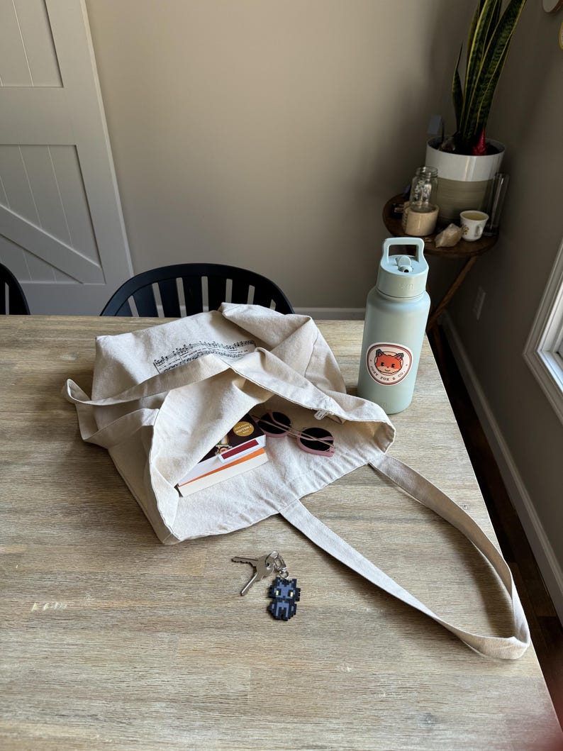 Puede incluir: Una bolsa tote de lona beige con un dise&ntilde;o impreso en negro, que contiene gafas de sol, un libro y otros art&iacute;culos. Una botella de agua azul claro con una pegatina de zorro y un llavero de gato pixelado tambi&eacute;n est&aacute;n sobre la mesa de madera.