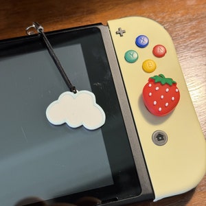 May include: A yellow Nintendo Switch console with a white cloud charm attached to the top. The console has a red and white strawberry-shaped button on the right joy-con.