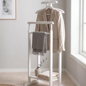 Puede incluir: Un galán de noche de madera blanca con una percha superior para un blazer beige, una barra horizontal que sostiene una bufanda gris con flecos y un estante inferior con un tejido doblado de color crema y un pequeño jarrón de cerámica con una rama verde.