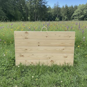 Puede incluir: Una jardinera rectangular de madera clara, con tablas horizontales y un asa de cuerda. La jardinera está situada en un campo de hierba verde y flores silvestres, con árboles y cielo azul de fondo.