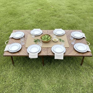 May include: A wooden picnic table set for eight people, with white plates, glasses, and linen napkins. A woven placemat sits in the center, holding a bowl of salad and small dishes. The table is set on a green lawn.