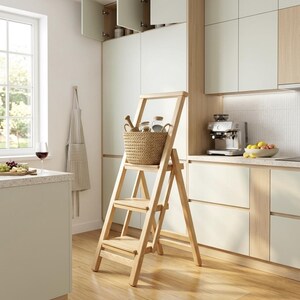 May include: A light-colored wooden three-step ladder with a tall handle, featuring a woven tan basket placed on the top step. The ladder has a sturdy frame with three horizontal treads and angled side supports, standing on a wooden floor.