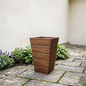 Puede incluir: Una jardinera alta y cuadrada de madera de color marrón oscuro. La jardinera tiene un diseño cónico, con listones de madera horizontales. Se encuentra sobre un patio de piedra, con plantas y flores en el fondo. La jardinera mide aproximadamente 76 cm de altura.