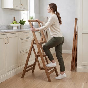Puede incluir: Una escalera de mano plegable de madera con tres peldaños, con un acabado de veta de madera marrón claro natural. La escalera tiene un diseño robusto en forma de A con un asa superior para mayor estabilidad, mostrada en una cocina con una unidad plegada.
