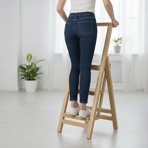 Op de afbeelding: Een lichtgekleurde houten trap met drie treden. De trap is voorzien van een hoge steunbeugel aan de bovenzijde en een inklapbaar frame. De constructie is gemaakt van natuurlijk hout met een gladde afwerking voor een stabiel platform.
