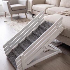 May include: A white wooden pet ramp with a slatted side design and a textured dark gray carpeted surface. The ramp features an adjustable folding support structure and is positioned at an incline to provide access to an elevated surface.