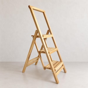Könnte beinhalten: Eine Trittleiter aus hellem Holz mit drei horizontalen Stufen und einem hohen, rechteckigen Griffrahmen an der Oberseite. Die Konstruktion wird durch Metallscharniere und Schrauben zusammengehalten und bietet ein stabiles, klappbares Design.