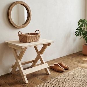 Op de afbeelding: Een lichtgekleurde houten klaptafel met een gevlochten mand erop. Een ronde houten spiegel hangt aan de muur boven de tafel. Bruine leren slippers staan op de vloer bij een geweven vloerkleed en een plant in een pot.