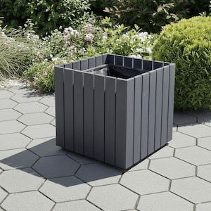 May include: A square, gray wooden planter box with vertical slats. The planter sits on a gray hexagonal paver patio, surrounded by greenery and flowers. The box is designed for outdoor use and has a dark interior.