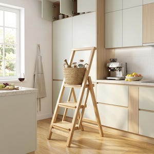 Puede incluir: Una escalera de mano de madera clara con tres peldaños y un asa alta en la parte superior. Una cesta tejida de color beige que contiene varios artículos de cocina descansa sobre el peldaño superior de la escalera, situada en un suelo de madera.