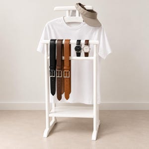 Könnte beinhalten: Ein weißer hölzerner Herrendiener mit einem weißen T-Shirt, einem beigen Visier, drei Ledergürteln in Schwarz, Dunkelbraun und Hellbraun sowie zwei Armbanduhren mit runden Zifferblättern, eine in Schwarz und eine in Weiß, auf dem Ständer.