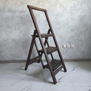 Könnte beinhalten: Eine dunkelbraune Holztrittleiter mit drei horizontalen Stufen und einem hohen, rechteckigen Griffrahmen an der Oberseite. Die Leiter ist mit stabilen Seitenholmen und Metallbeschlägen konstruiert, die die klappbaren Rahmenteile verbinden.