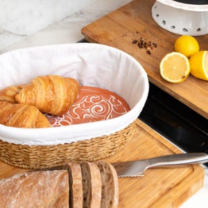 May include: A woven wicker bread basket with a white liner holds golden croissants. A terracotta plate with a white swirl design sits inside. Sliced bread and a knife rest on a wooden cutting board, with lemons and a white cake stand in the background.
