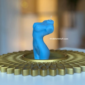 May include: A blue candle shaped like a human torso, sitting on a gold decorative stand. The candle is lit and the wick is visible. The text "sculpturesstuff.com" is visible on the stand.
