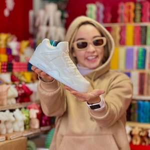 May include: A white sneaker candle, a popular gift for sneakerheads and candle enthusiasts. The candle is shaped like a classic basketball shoe and is being held by a person wearing a beige hoodie.