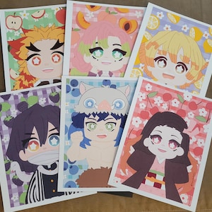 May include: A collection of six colorful art prints featuring anime-style characters. Each print showcases a different character with unique hair and eye colors, set against backgrounds with fruit and floral patterns. The prints are on a brown surface.