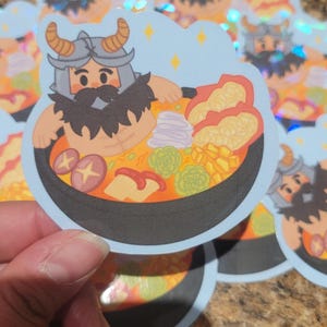 May include: A sticker featuring a cartoon illustration of a bearded warrior in a helmet, relaxing in a bowl of steaming hot soup. The soup is filled with various vegetables and noodles. The sticker has a holographic finish.