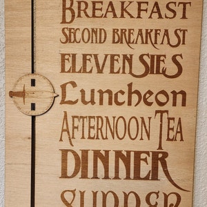 Op de afbeelding: Houten bord met een schuifregelaar die "Now Serving" aangeeft en verschillende maaltijden opnoemt, waaronder "Breakfast", "Second Breakfast", "Elevensies", "Luncheon", "Afternoon Tea", "Dinner" en "Supper".