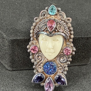 May include: An ornate silver-toned pendant featuring a carved face with a serene expression. The pendant is adorned with various colorful gemstones, including pink, blue, and purple, set within a detailed, swirling design. The overall aesthetic is reminiscent of vintage jewelry.