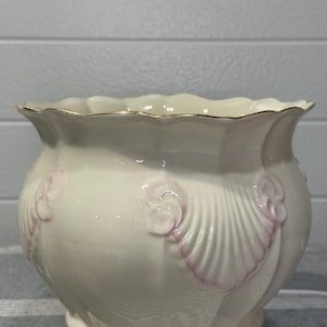 May include: Cream-colored ceramic planter with a scalloped rim and embossed shell designs. The planter has a gold trim and pink accents on the shell details. A decorative container for plants.