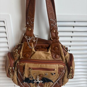 May include: A brown and tan leather handbag with multiple pockets and zippers. The bag is decorated with silver chains, charms, and safety pins. A black bat wing and cross charm are on the front.