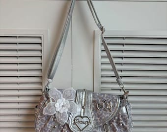 Peut inclure: Un sac à main argenté et gris avec un motif léopard. Le sac présente une grande fleur blanche, un emblème en forme de cœur avec le mot "Poarapy" et des accents argentés. Une bandoulière argentée est attachée au sac.